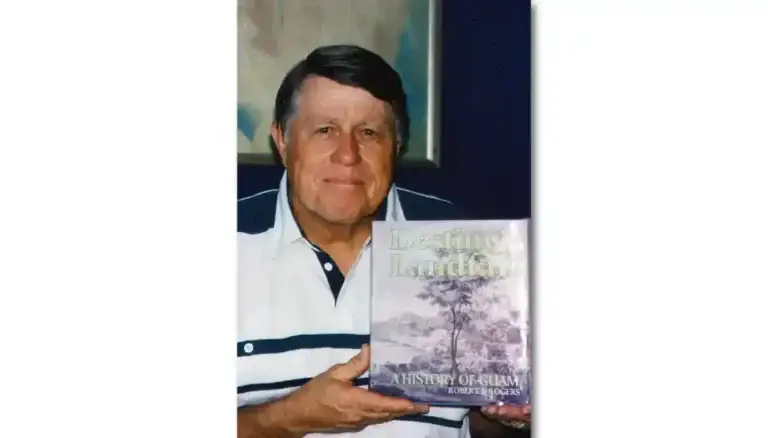 Portrait of an older man in a white and navy polo holding a book titled 'A History of Guam' indoors, with framed artwork in the background.