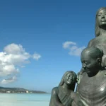 Bronze statue of a mother with two children by the seaside under a blue sky.