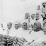 Group of women in white robes and headscarves seated in stadium bleachers, with a man wearing a hat in the background.