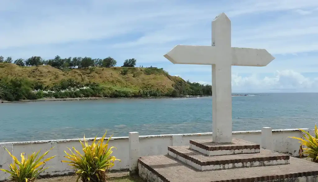 The San Dionisio Church cross located across the street. Behind are the Humåtak/Umatac Cemetery and Fort Soledad. Photo by George Miller, 2014.