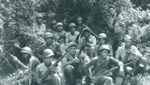 Group of soldiers with rifles posing in a jungle setting, with one woman standing at the back right side of the group.