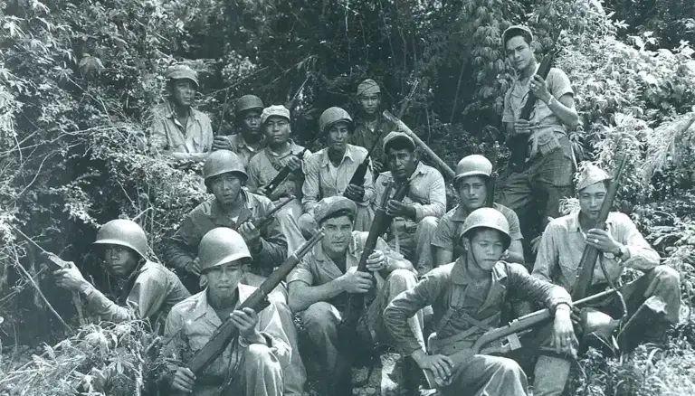 Group of soldiers with rifles posing in a jungle setting, with one woman standing at the back right side of the group.