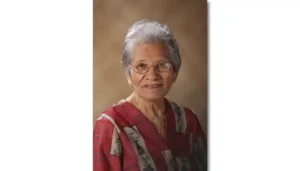 Portrait of an elderly woman with short gray hair and glasses wearing a red patterned blouse, smiling softly.