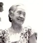 Older woman smiling, wearing a floral dress, with leaves in the foreground in a black-and-white photo