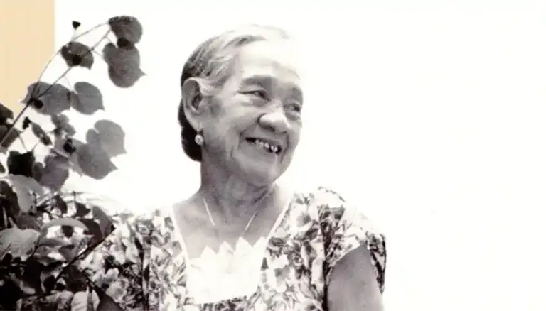 Older woman smiling, wearing a floral dress, with leaves in the foreground in a black-and-white photo