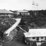 Black-and-white photo of a hillside campus with low, wraparound buildings, a winding dirt road, and two tall water towers in the distance.