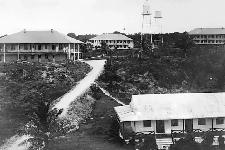 Black-and-white photo of a hillside campus with low, wraparound buildings, a winding dirt road, and two tall water towers in the distance.