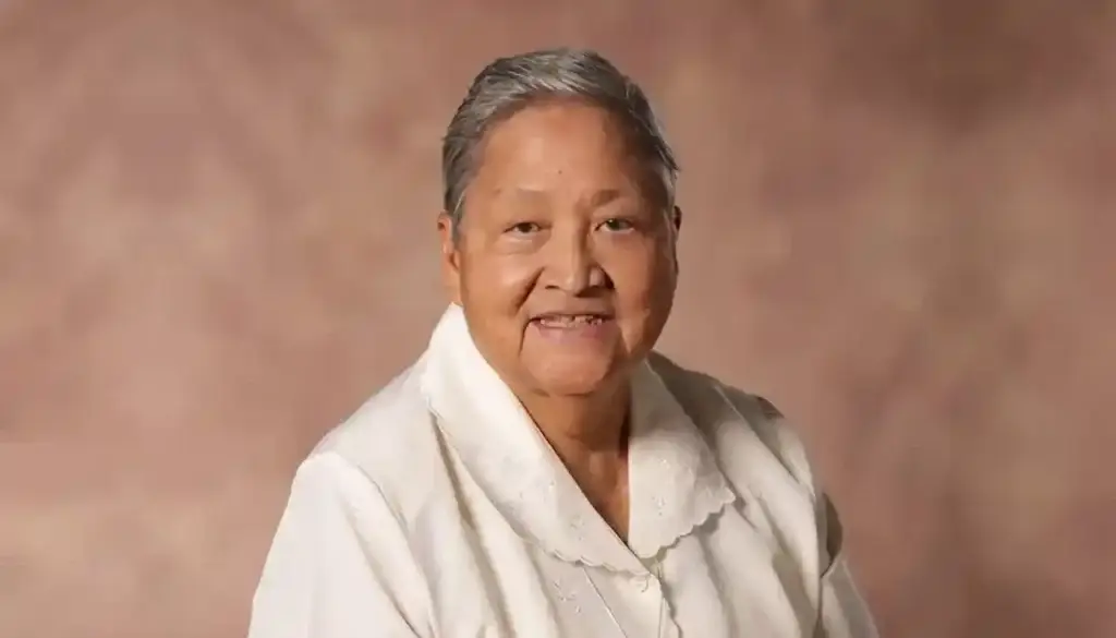 Smiling elderly woman with short gray hair in a white blouse against a warm brown studio backdrop.