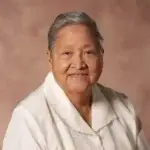 Smiling elderly woman with short gray hair in a white blouse against a warm brown studio backdrop.