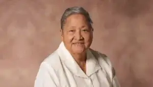 Smiling elderly woman with short gray hair in a white blouse against a warm brown studio backdrop.