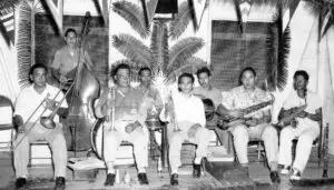 A black-and-white photo of a nine-person band posing with brass, string, and percussion instruments, including a double bass, trumpets, trombone, guitar, and a saxophone in a tropical-themed backdrop.