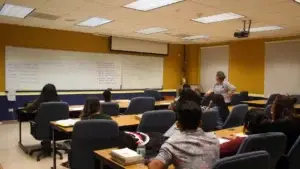 Instructor in a striped shirt stands at the front of a classroom, guiding students seated at long desks.